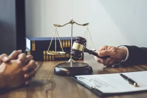 Dłoń trzymająca młotek sędziowski, waga sprawiedliwości i akta na drewnianym stole.