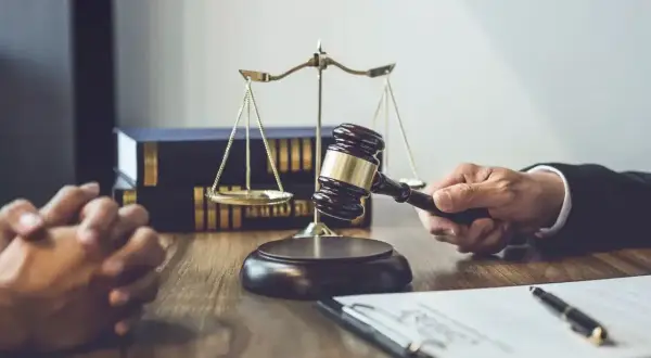 Dłoń trzymająca młotek sędziowski, waga sprawiedliwości i akta na drewnianym stole.