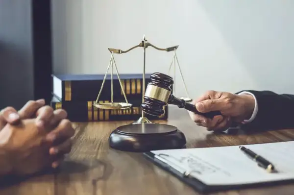 Dłoń trzymająca młotek sędziowski, waga sprawiedliwości i akta na drewnianym stole.